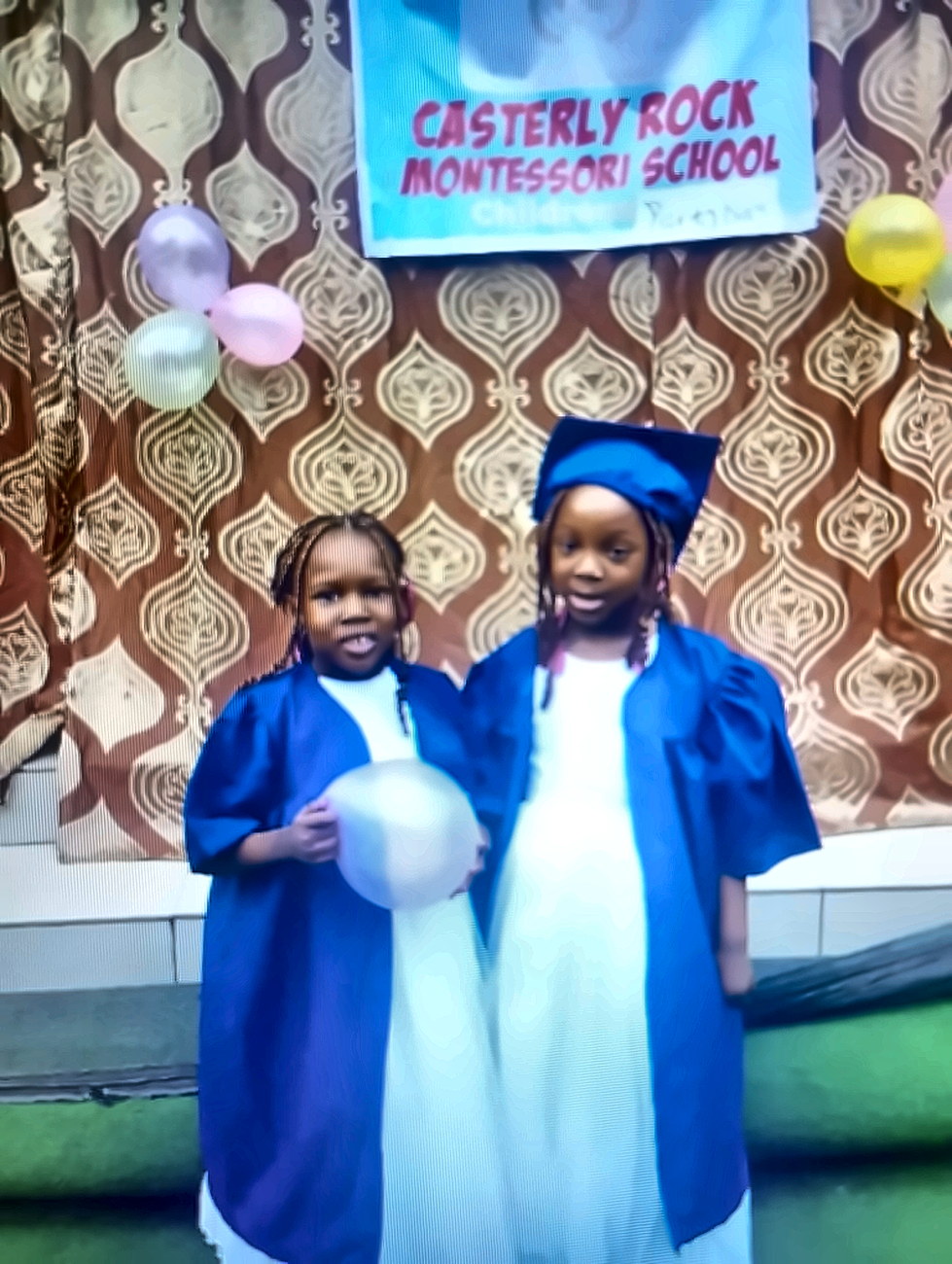 Two pupils in blue graduation gowns beneath the Casterly Rock Children&rsquo;s Party Day banner